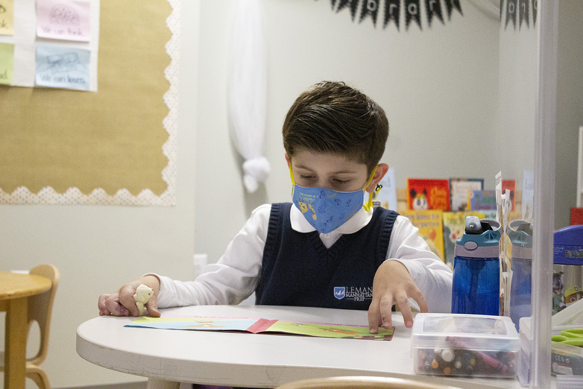 A boy sits at a desk reading with a mask on