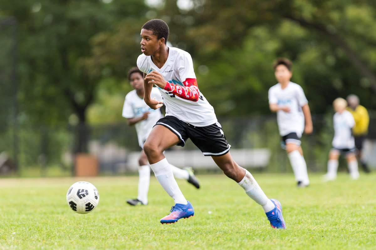 A student on the Middle School soccer team runs, kicking a ball in front of him. He is in a white and blue uniform.